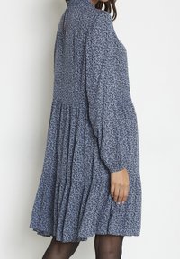 Mujer con un vestido azul de manga larga y holgado, con pequeños estampados florales negros, de pie frente a un fondo blanco.
