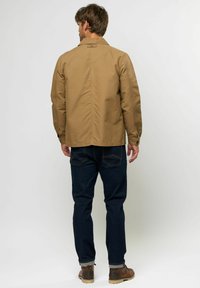 Chaqueta caqui con costuras visibles y espalda ventilada, combinada con jeans de denim oscuro, puños enrollados y botas marrones de cordones.
