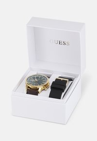 Orologio tono oro con quadrante nero e cinturino in pelle marrone, abbinato a un cinturino in gomma nero, esposto in una scatola bianca con la scritta "GUESS".