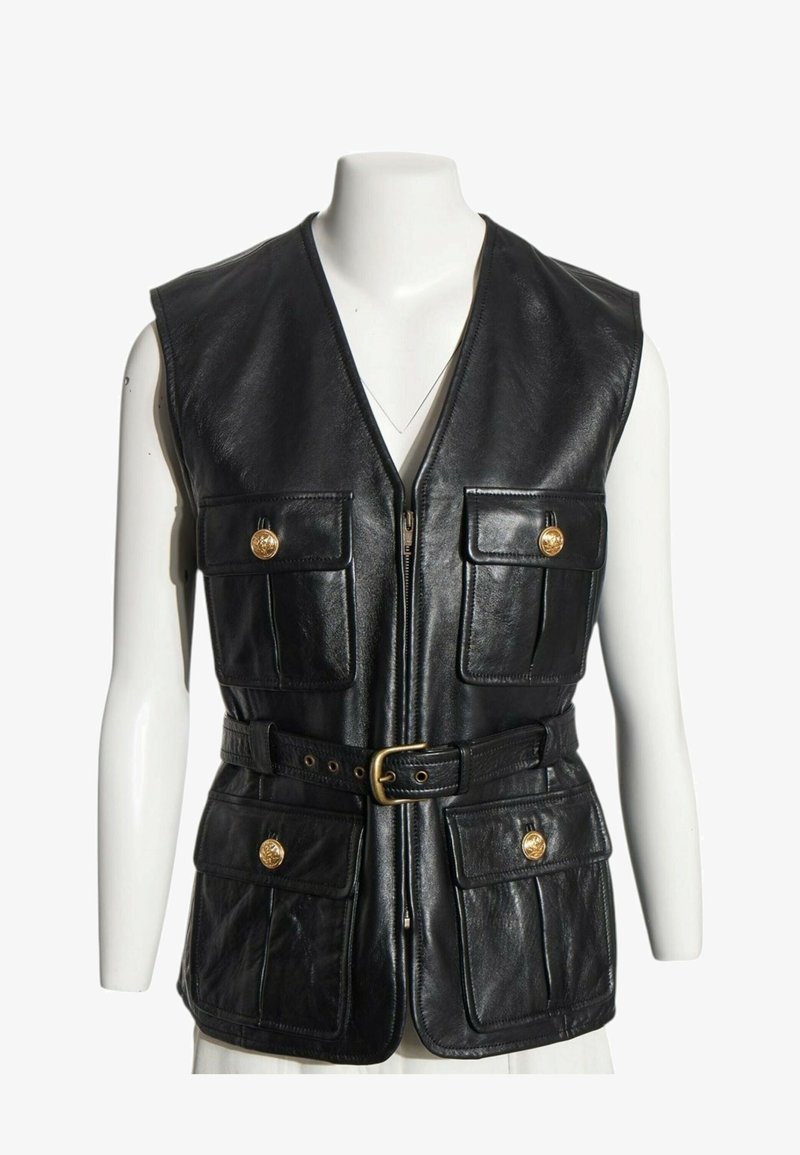 Veste sans manches en cuir noir avec fermeture éclair sur le devant, quatre poches à rabat avec boutons dorés, et une ceinture avec boucle dorée à la taille sur un mannequin blanc.