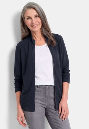 Lachende vrouw met lang grijs haar draagt een zwart ritsjack over een wit overhemd en grijze broek tegen een effen lichte achtergrond.