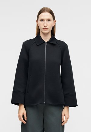 Femme portant une veste noire zippée avec des manches larges et un col, debout devant un fond uni gris clair.