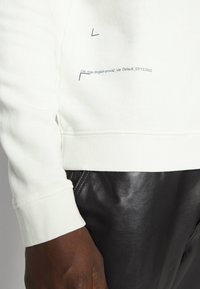 Felpa bianca con polsini a coste, caratterizzata da testo stampato blu e design del logo. Pantaloni in pelle nera parzialmente visibili sotto la felpa.