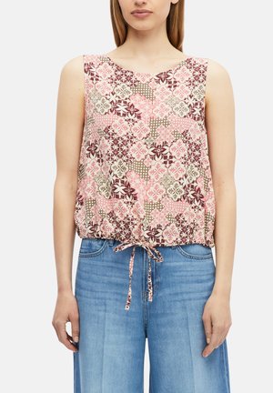 Frau trägt ärmelloses pinkes Oberteil mit floralem Muster und Bindeband an der Taille sowie hochgeschnittene blaue Jeans vor weißem Hintergrund.
