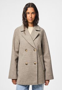 Cappotto beige doppiopetto con grandi bottoni dorati, ampi rever e maniche diritte. Indossato sopra un top chiaro e jeans di denim.