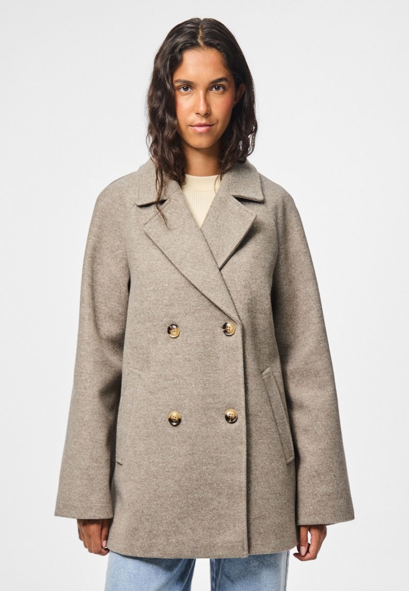 Cappotto beige doppiopetto con grandi bottoni dorati, ampi rever e maniche diritte. Indossato sopra un top chiaro e jeans di denim.