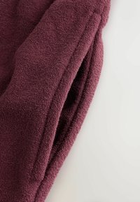 Plüschiges bordeauxfarbenes Gewebe, weiche Textur mit einem geschlungenen Design. Verfügt über eine seitliche Nahttasche für den praktischen Gebrauch. Glatte Oberfläche, leicht und gemütlich.