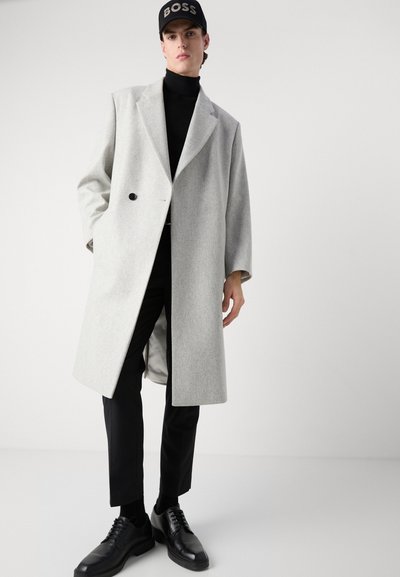 Manteau en laine gris clair avec un design double boutonnage, détails de boutons noirs, associé à un col roulé noir et un pantalon sombre, chaussures noires.