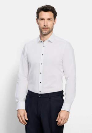 Mann trägt ein eng anliegendes weißes Hemd mit langen Ärmeln, schwarzen Knöpfen und dunkle, maßgeschneiderte Hose vor einfarbigem Hintergrund.