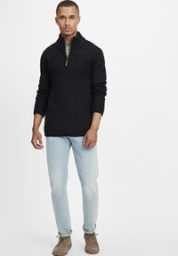 Strikket sort sweater med høj krave, halvlukning, tekstureret overdel og farvestræk rundt om, parret med lyseblå jeans.