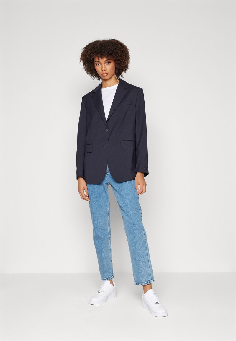 Blazer navy avec revers en notch, deux poches avant et une fermeture à un bouton, porté sur un t-shirt blanc et associé à un jean bleu clair.