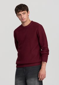 Maglione lavorato a maglia bordeaux con scollo rotondo, tessuto testurizzato e polsini e orlo a coste, abbinato a jeans scuri.