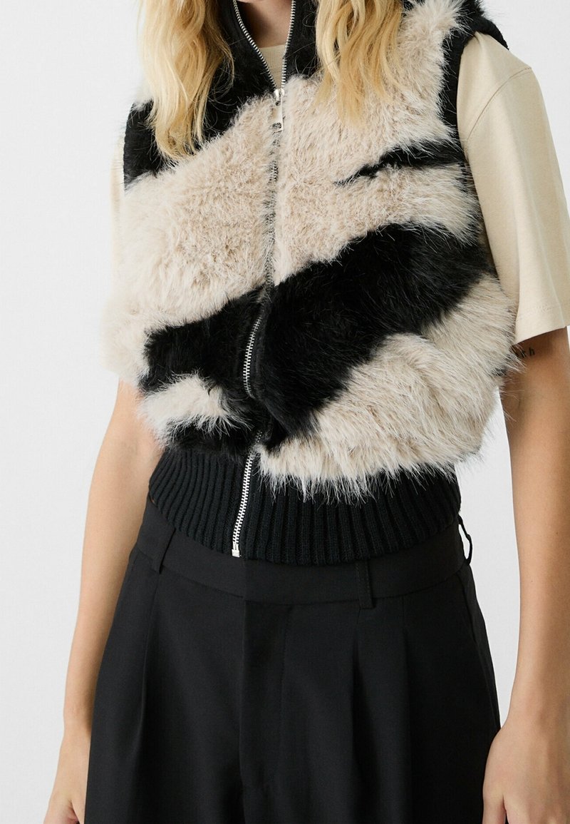 Femme portant un gilet sans manches en fausse fourrure noire et beige avec fermeture éclair, par-dessus une chemise beige à manches courtes et un pantalon noir taille haute.