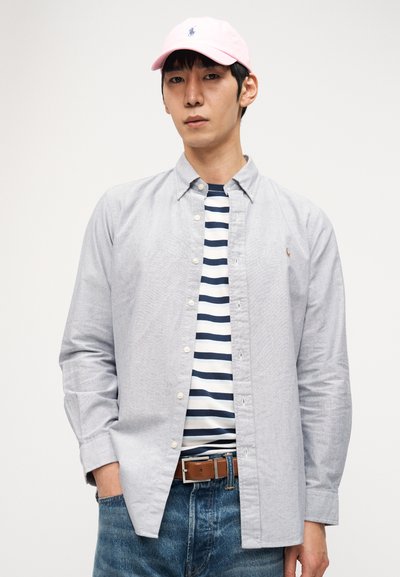 Άνδρας που φοράει ανοιχτό γκρι πουκάμισο με κουμπιά πάνω από μπλε σκούρο και λευκό ριγέ μπλουζάκι, ανοιχτό ροζ καπέλο, καφέ ζώνη και μπλε τζιν, με λευκό φόντο.