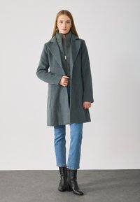 Cappotto in misto lana grigio con colletto, tasche frontali e vestibilità morbida, abbinato a jeans blu a gamba dritta e stivaletti neri.