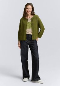 Green ribbed cardigan with button closure, layered over a beige graphic t-shirt, paired with dark flared jeans and white sneakers.