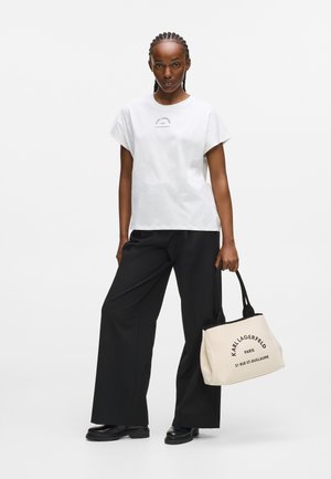 White cotton t-shirt with short sleeves, paired with loose black trousers. Carrying a beige tote bag with black accents and printed text.