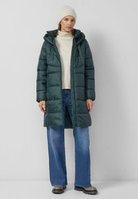 Ciemnozielona ocieplana kurtka z kapturem, z zamek błyskawicznym i sznurkami do regulacji. Zestawiona z beżowym swetrem i spodniami z szerokimi nogawkami.