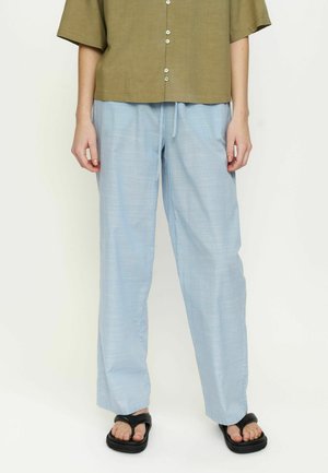 Person iført lyseblå bukser med brede ben, olivengrøn kortærmet skjorte med knapper og sorte plateformesandaler, stående mod en ensfarvet baggrund.