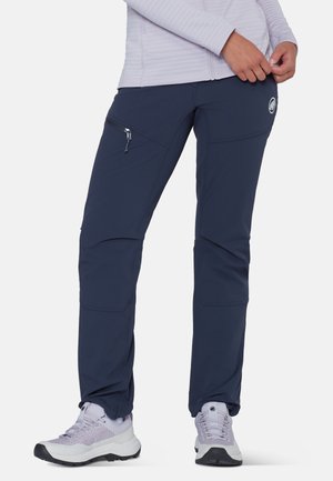 Persona che indossa pantaloni da trekking blu navy con cerniera e scarpe da trekking grigio chiaro, in piedi contro uno sfondo semplice.