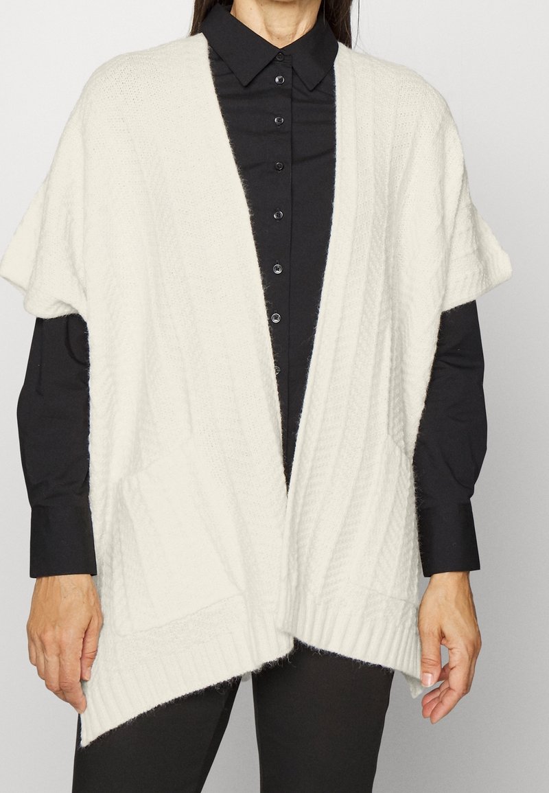 Femme portant un gilet sans manches en tricot blanc sur une chemise noire boutonnée et un pantalon noir, debout devant un fond clair.