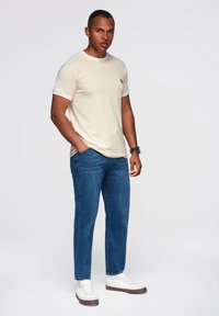 Beige tričko s krátkým rukávem a bílým lemem, spárované s modrými džíny a bílými teniskami. Oblečení má volný střih a jednoduchý design.