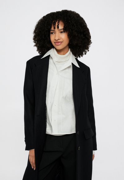 Must pikk mantel märgiga kraega, millele on lisatud valge nööbitav särk ja tekstuuriga kaelasall, taustaks on lihtne sein.