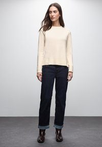 Pullover lavorato a maglia color beige chiaro con orlo a coste, abbinato a jeans blu scuro con risvolto in fondo e stivali neri alla caviglia.
