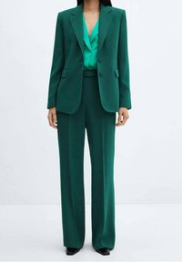 Abito verde su misura con blazer strutturato e revers, abbinato a pantaloni a gamba larga. Sotto, una camicia di raso che aggiunge una texture a contrasto.