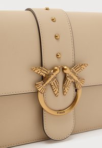 Τσάντα χειρός από μπεζ δέρμα με χρυσό κυκλικό κούμπωμα διακοσμημένο με δύο πουλιά. Λεπτή υφή με διακοσμητικά καρφιά κατά μήκος της πλαϊνής πτέρυγας.