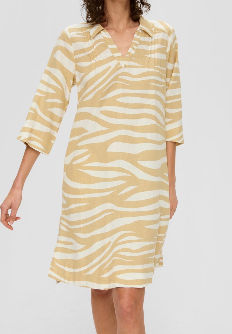 Femme portant une robe beige et blanche à rayures zébrées, longueur genou, avec des manches trois-quarts et un col en V.