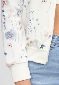 Veste bomber florale présentant des fleurs bleu pastel et rose sur un fond blanc, avec des poignets en côte blancs et une fermeture éclair à l'avant.