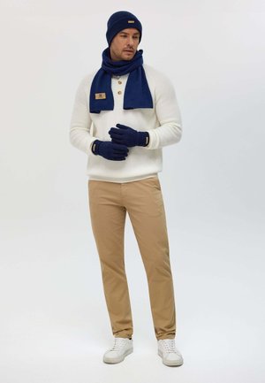 Weißer Strickpullover mit drei Knöpfen, marineblauer Schal, Handschuhe und Mütze. Tanfarbene Hose und weiße Sneaker vervollständigen das Outfit. Einfaches und modernes Design.