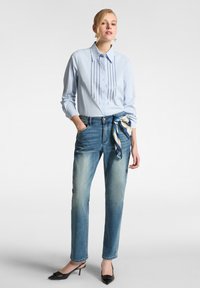 Camicia azzurra con bottoni e dettagli verticali in perline, abbinata a jeans in denim azzurri con una sciarpa decorativa annodata in vita. Scarpe nere con tacco.