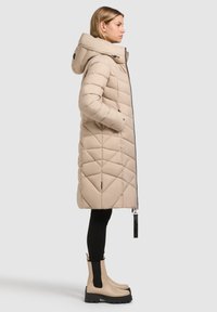 Palton puffer bej, lungime până la genunchi, cu glugă, având modele lasă de tip diamant și fermoar negru. Purtați cu colanți negri și boots.