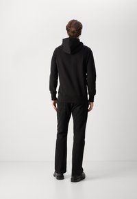 Felpa nera con cappuccio regolabile, tessuto testurizzato, polsini e orlo a coste, indossata con jeans neri e scarpe nere. Vista posteriore.