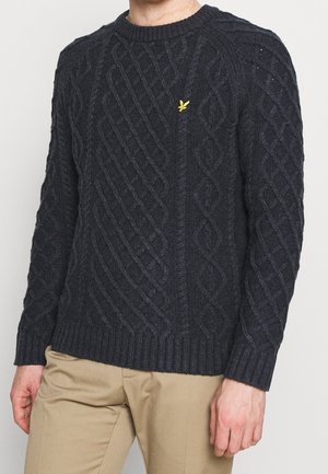 Homme portant un pull en maille torsadée gris foncé avec un petit logo oiseau jaune sur la poitrine et un pantalon beige, debout devant un fond uni.