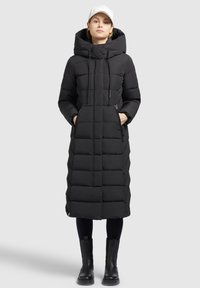 Sort polstret lang frakke med hætte, der har lynlåslommer, et quiltet mønster og en lige silhuet. Båret med sorte støvler.