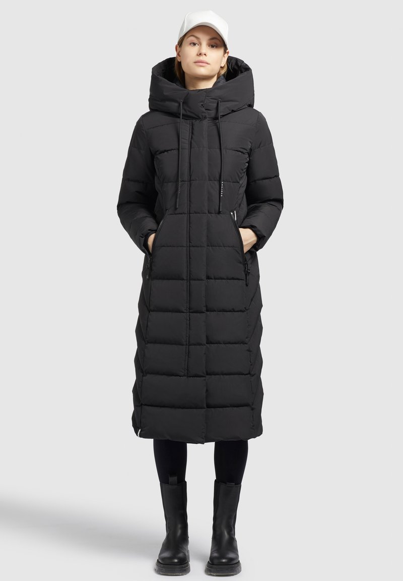 Sort polstret lang frakke med hætte, der har lynlåslommer, et quiltet mønster og en lige silhuet. Båret med sorte støvler.