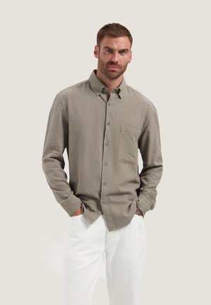 Mann trägt ein beige langärmliges Hemd mit Brusttasche und weiße Hose, steht mit den Händen in den Taschen vor schlichtem Hintergrund.