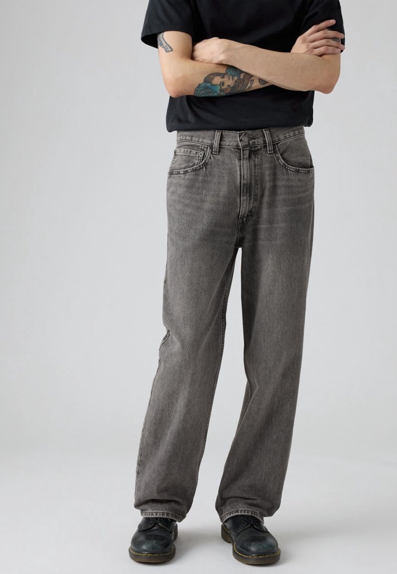 Personne aux bras croisés portant une chemise noire, un jean gris ample, des chaussures noires et des tatouages visibles sur les bras, debout sur un fond gris uni.