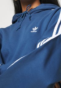 Granatowa bluza z kapturem Adidas z regulowanym sznurkiem, białymi trzema paskami na rękawach oraz małym białym logo na piersi.