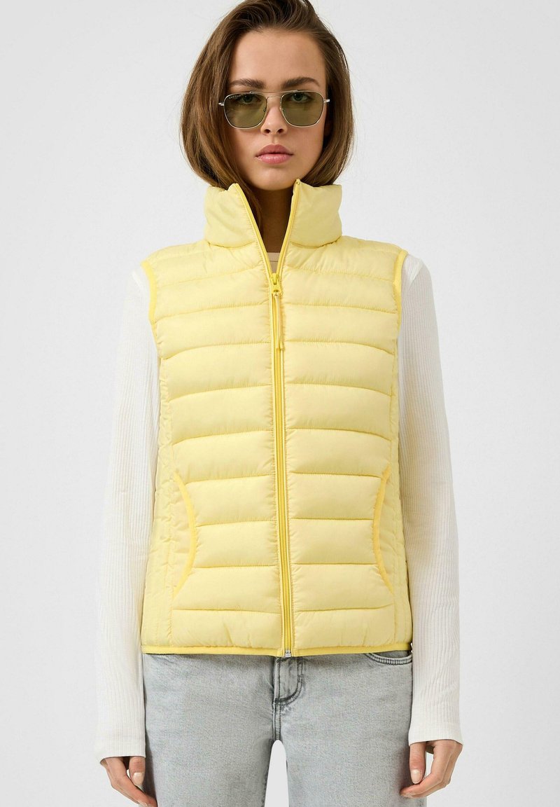 Gelbe Steppweste mit hohem Kragen, horizontaler Steppung, Reißverschluss und zwei seitlichen Taschen, kombiniert mit einem weißen Langarmshirt.