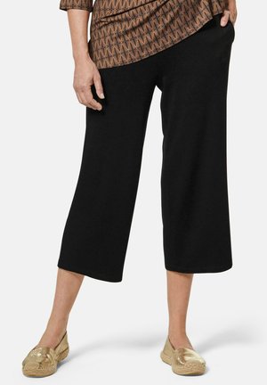 Schwarze, verkürzte Hose mit lockerer Passform, aus weichem Stoff gefertigt. Kombiniert mit goldenen Espadrilles. Geometrisch gemustertes Oberteil sichtbar.