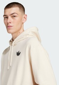 Cream hoodie made of soft fabric with a drawstring, featuring a black stylized logo on the left chest area.