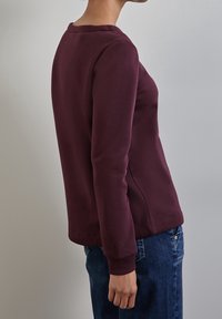 Sudadera de cuello redondo en color marrón burdeos con mangas largas y puños acanalados. Textura suave con un diseño ajustado, combinada con jeans de mezclilla azules.