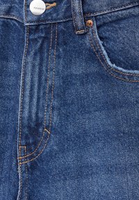 Gros plan d'un jean en denim bleu montrant la poche avant, la passants de ceinture, la couture orange et un bouton argenté avec gravure de la marque.