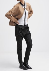 Osoba w beżowym kardiganie na białej koszuli z zapięciem na guziki i czarnymi wstawkami, w czarnych spódnicach z wysokim stanem i czarnych botkach za kostkę, stoi z rękami na biodrach.