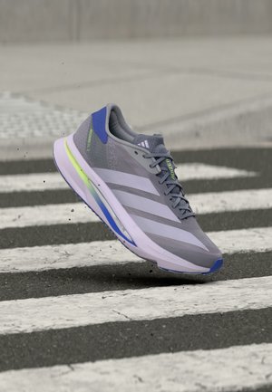 Γκρι παπούτσι τρεξίματος Adidas με μπλε και πράσινες λεπτομέρειες, στον αέρα πάνω από διάβαση πεζών με ασπρόμαυρες ρίγες σε άσφαλτο.
