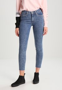 Jeans ajustados de cintura alta en denim azul claro con un acabado desgastado y dobladillo deshilachado. Combinados con botas negras de tobillo y una blusa de color rosa pastel.